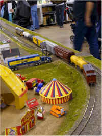 The American Flyer semi truck still waits as an American Models Fairbanks Morse "Train Master" H-24-66 locomotive on the point of a freight train hiballs past on the inner mainline.