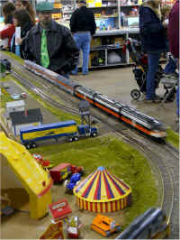 An American Flyer semi truck waits as an American Models Alco PA-PB-PA on the point of a Budd streamliner consist passes on the outside mainline track.