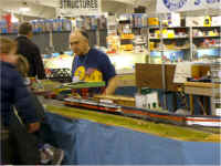 Roger Delthony takes a turn monitoring the accessories as an American Models Alco PA-PB-PA in New Haven livery on the point of a Budd streamliner consist hiballs down the accessories side of the modular layout.